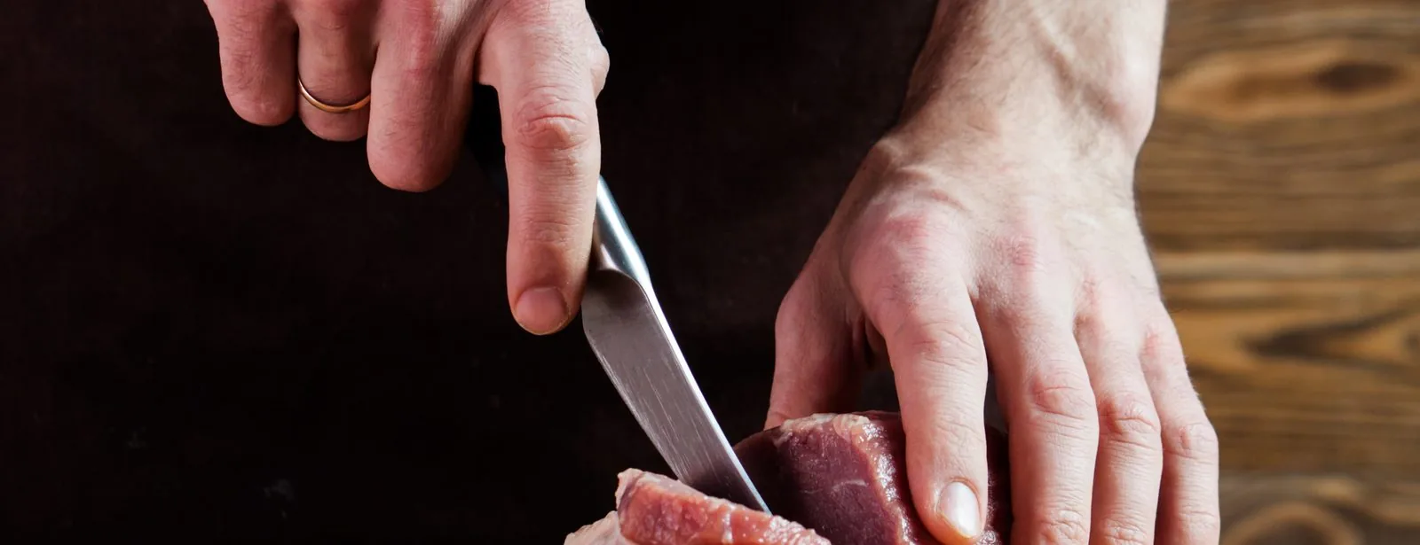 Hands slicing raw meat with a steel knife on a dark prep surface