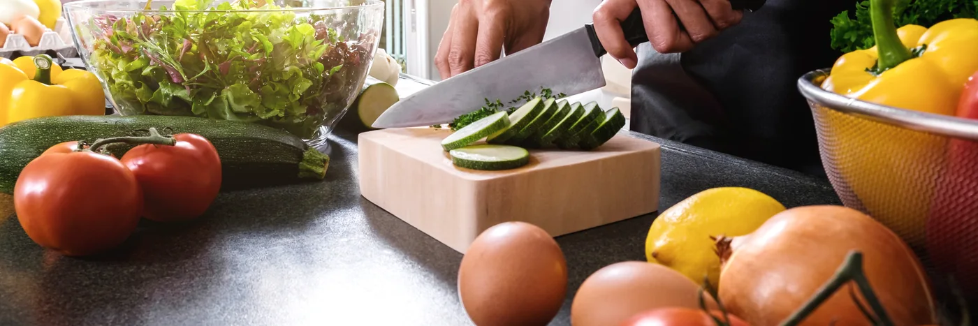 Hands slicing cucumber on a board surrounded by fresh vegetables.