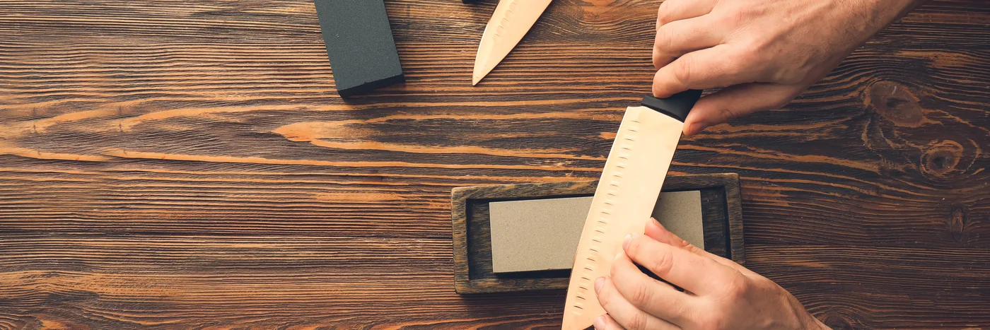 Hands sharpening a kitchen knife on a whetstone on a wooden bench.
